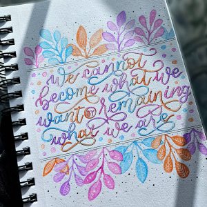Hand‑lettered quote reading “We cannot become what we want by remaining what we are” in shades of blue, green, orange, and pink