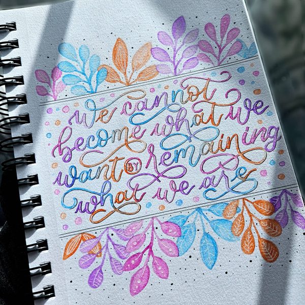 Hand‑lettered quote reading “We cannot become what we want by remaining what we are” in shades of blue, green, orange, and pink