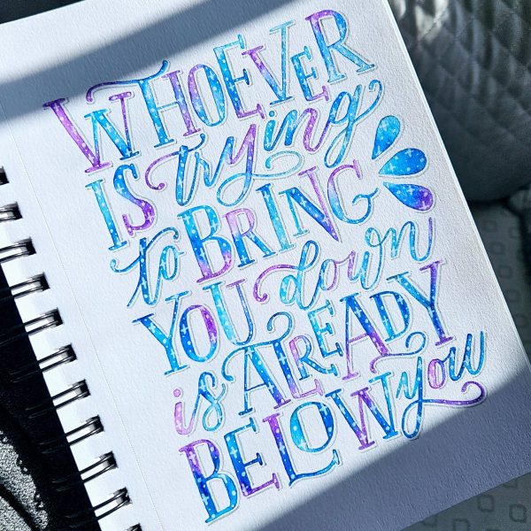 Hand‑lettered quote reading “Whoever is trying to bring you down is already below you” painted in blue and purple
