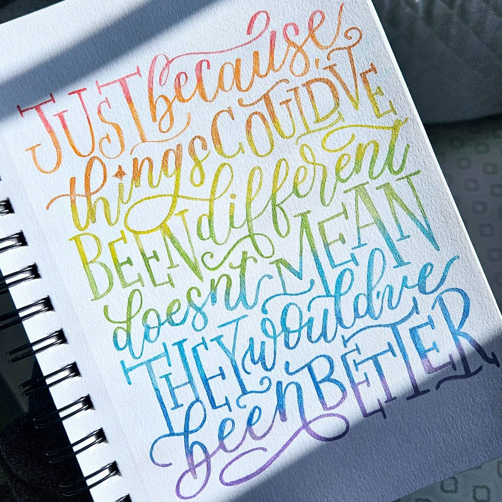 Hand‑lettered quote reading “Just because things could’ve been different doesn’t mean they would’ve been better” painted in rainbow pearlescent watercolors.