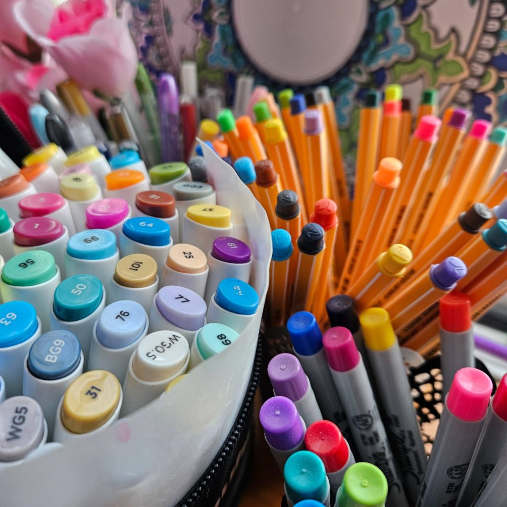 A close-up of various art supplies: a pouch that contains alcohol markers, a cup that contains Stabilo fineliners, and a cup with Sharpies